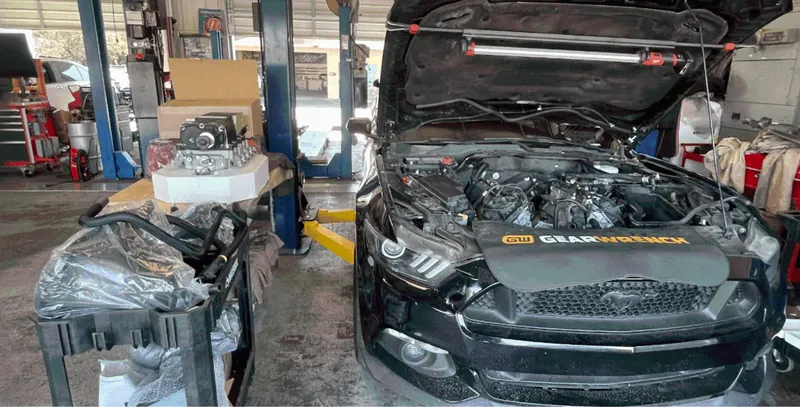 Inside the Tulare Automotive Repair bay: a black Mustang with its hood up and a fender cover over the front, new parts boxed on the bench beside it, blue two-post lifts behind.