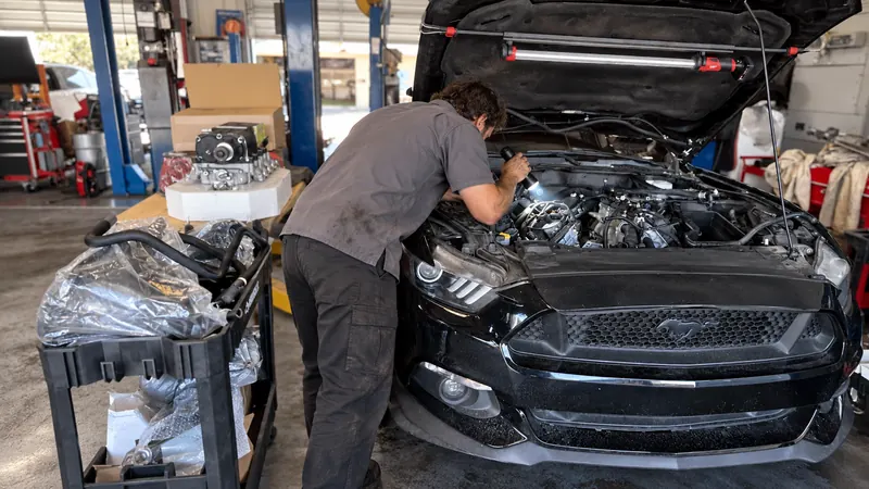 A technician leans into the Mustang's engine bay with a flashlight, parts boxes on the bench behind him.